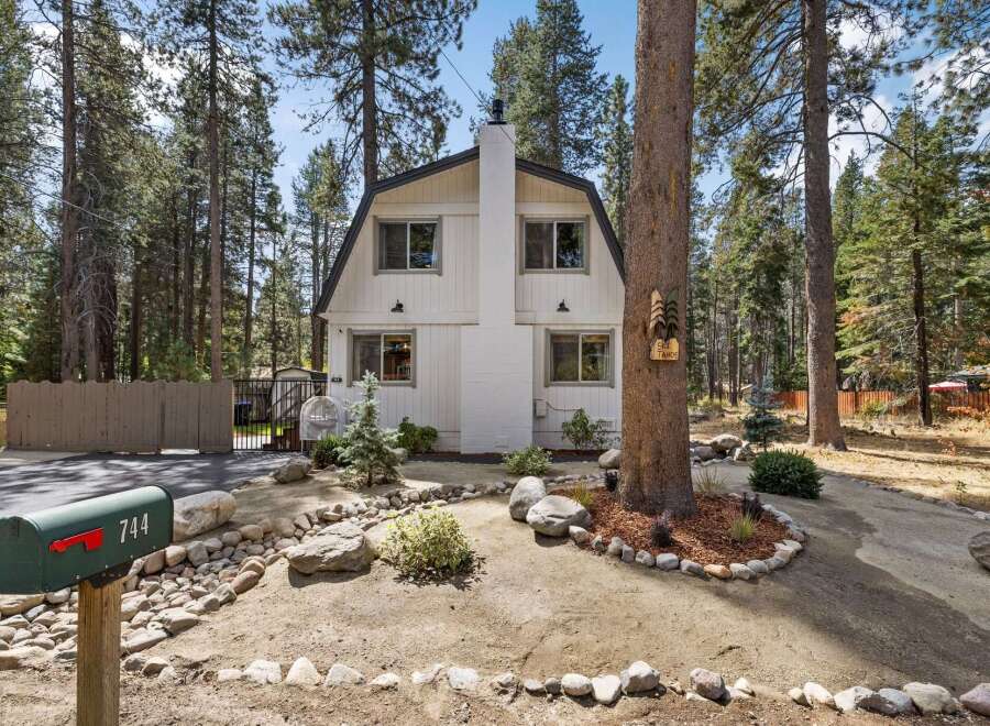 Charming cabin nestled among tall pine trees, featuring a distinctive gambrel roof and a prominent white brick chimney.