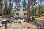 Charming cabin nestled among tall pine trees, featuring a distinctive gambrel roof and a prominent white brick chimney.