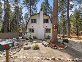 Charming cabin nestled among tall pine trees, featuring a distinctive gambrel roof and a prominent white brick chimney.