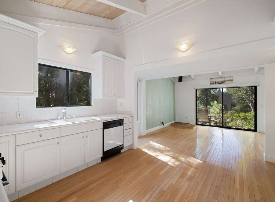 Bright and airy kitchen with white cabinetry and a spacious sink, opening up to a sun-drenched living area with large windows offering serene views of the surrounding greenery.