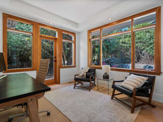 This home office boasts abundant natural light streaming through large wooden-framed windows, offering serene views of lush greenery.
