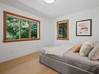 This bedroom offers a serene retreat with abundant natural light streaming through large, wood-trimmed windows overlooking lush greenery.