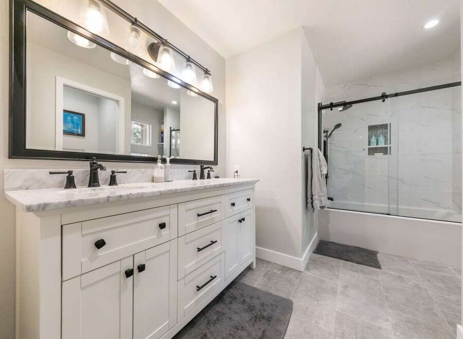 This modern bathroom features a spacious white double vanity with marble countertops and black hardware, complemented by a large black-framed mirror with an industrial-style light fixture.