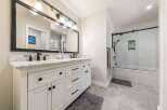 This modern bathroom features a spacious white double vanity with marble countertops and black hardware, complemented by a large black-framed mirror with an industrial-style light fixture.
