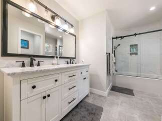 This modern bathroom features a spacious white double vanity with marble countertops and black hardware, complemented by a large black-framed mirror with an industrial-style light fixture.