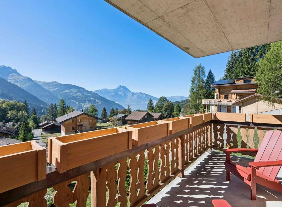 Enjoy breathtaking mountain views from this charming balcony, complete with wooden planters and a comfortable red Adirondack chair.