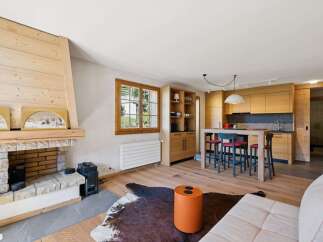 Experience modern alpine living in this beautifully appointed open-plan space, featuring a cozy stone fireplace and a sleek, wood-paneled kitchen.