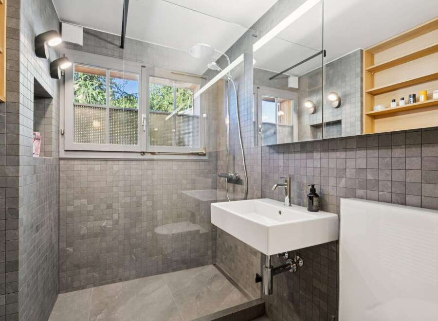 This modern bathroom features a sleek design with grey tiled walls and floors, complemented by a floating white sink and a glass-enclosed shower.