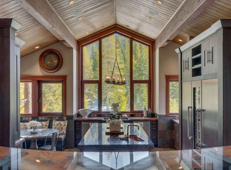 Experience luxury mountain living in this stunning kitchen featuring a grand vaulted ceiling with exposed beams and a massive A-frame window showcasing breathtaking forest views.