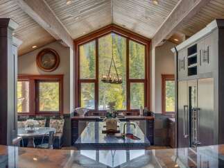 Experience luxury mountain living in this stunning kitchen featuring a grand vaulted ceiling with exposed beams and a massive A-frame window showcasing breathtaking forest views.