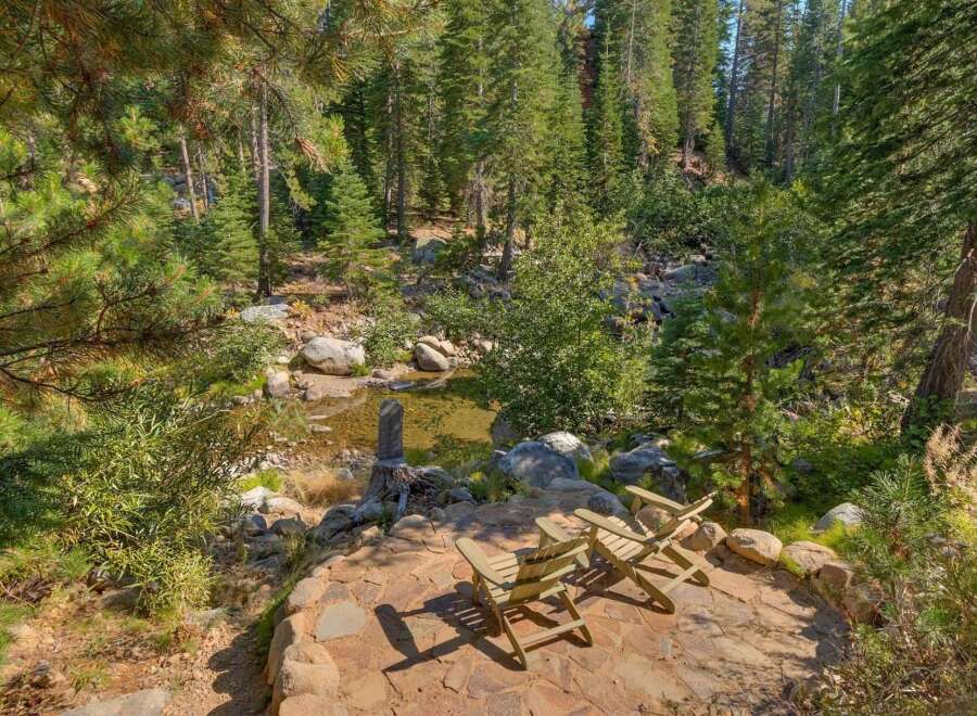 Escape to this serene mountain retreat featuring a tranquil creek and a charming stone patio with Adirondack chairs, perfect for enjoying the surrounding pine forest.