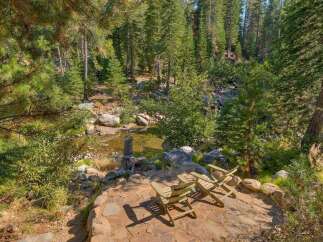 Escape to this serene mountain retreat featuring a tranquil creek and a charming stone patio with Adirondack chairs, perfect for enjoying the surrounding pine forest.