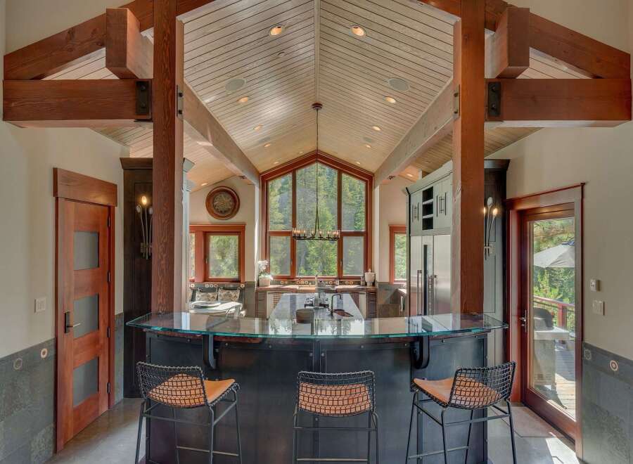 Experience luxury mountain living in this stunning kitchen, featuring a vaulted wood-paneled ceiling with exposed beams and a dramatic floor-to-ceiling window overlooking a lush forest.