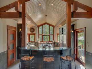 Experience luxury mountain living in this stunning kitchen, featuring a vaulted wood-paneled ceiling with exposed beams and a dramatic floor-to-ceiling window overlooking a lush forest.