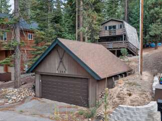 Nestled amidst tall pine trees, this charming mountain property features a rustic wooden exterior and a spacious two-car garage adorned with decorative skis and the house number 1369.