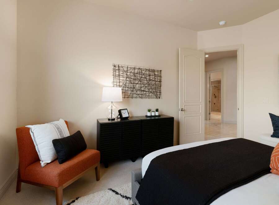 This inviting bedroom features a stylish orange accent chair with decorative pillows, complemented by a sleek black dresser adorned with a modern lamp and minimalist decor.