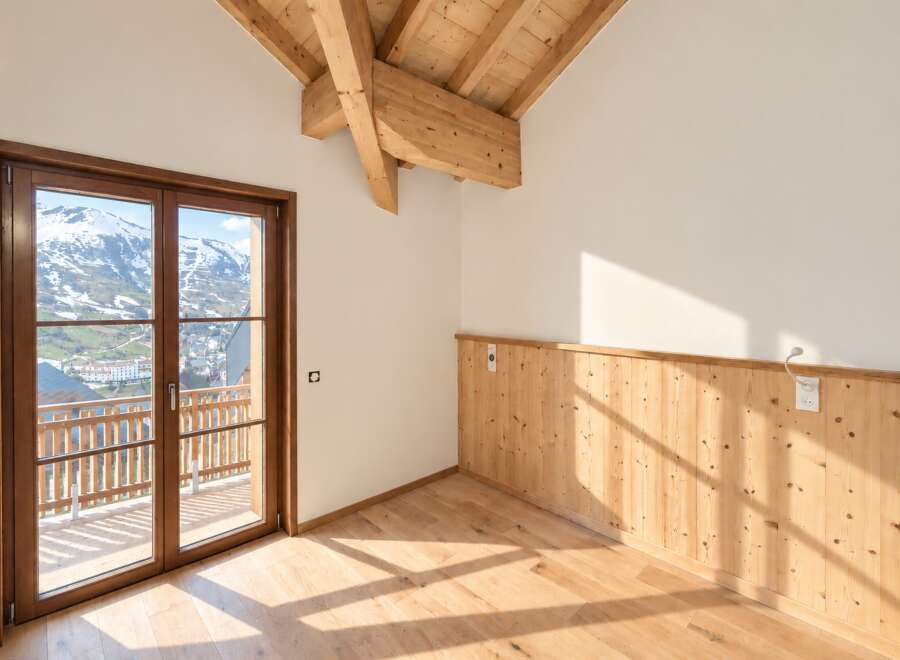 Experience breathtaking mountain views from this stunning property, featuring a spacious room with a wooden-beamed vaulted ceiling and a large glass door opening to a balcony.