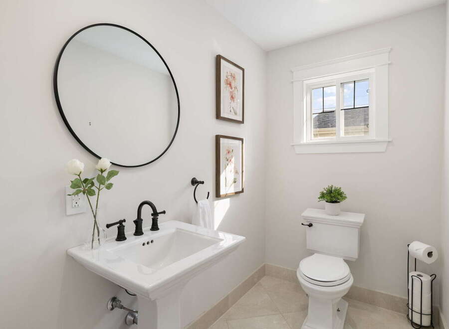 This bright and airy bathroom features a stylish pedestal sink with elegant black fixtures, complemented by a large round mirror and decorative floral artwork.