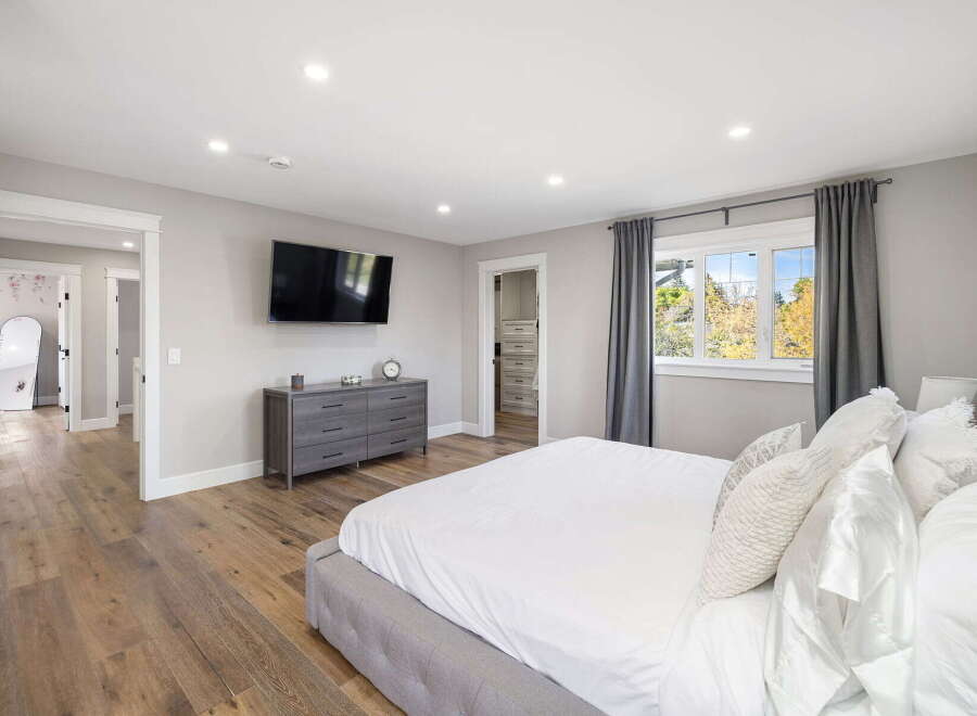 This spacious master bedroom features a large king-size bed with plush white bedding, a wall-mounted television, and a modern gray dresser.