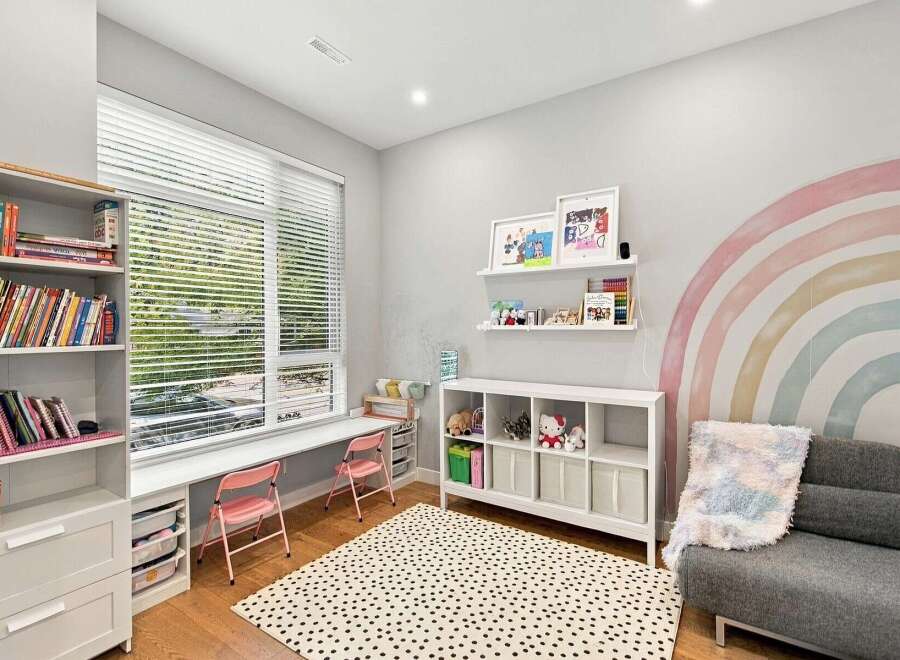This charming children's room features a large window with blinds, a built-in desk area with pink chairs, and a white shelving unit filled with toys and books.