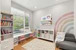 This charming children's room features a large window with blinds, a built-in desk area with pink chairs, and a white shelving unit filled with toys and books.
