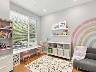 This charming children's room features a large window with blinds, a built-in desk area with pink chairs, and a white shelving unit filled with toys and books.