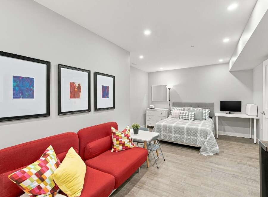 Discover this charming studio apartment featuring a vibrant red sofa and a comfortable queen-sized bed adorned with stylish grey and white bedding.