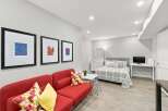 Discover this charming studio apartment featuring a vibrant red sofa and a comfortable queen-sized bed adorned with stylish grey and white bedding.