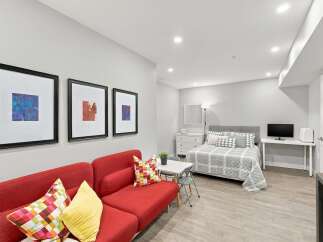 Discover this charming studio apartment featuring a vibrant red sofa and a comfortable queen-sized bed adorned with stylish grey and white bedding.