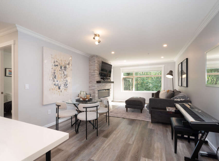Discover modern living in this beautifully appointed open-concept space, featuring a stylish dining area with a glass-top table and chic chairs, adjacent to a cozy living room with a stone-accented fireplace and a large window overlooking lush greenery.
