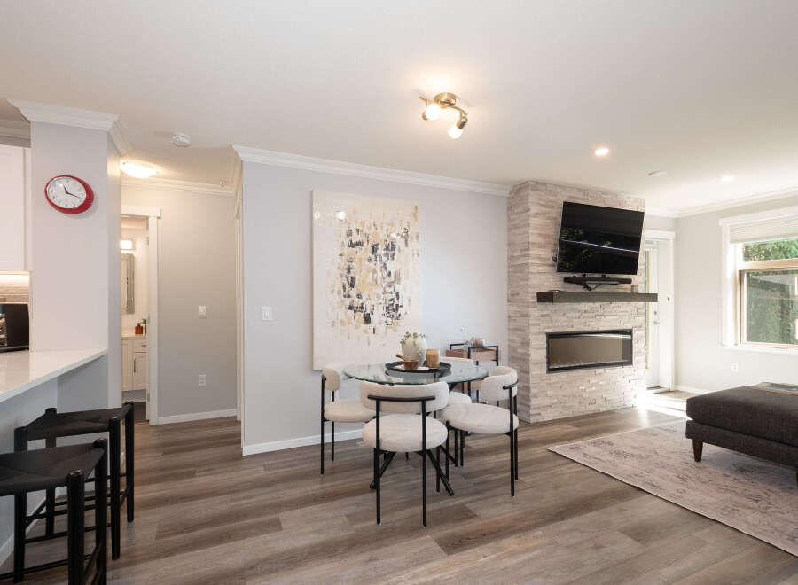 Experience modern living in this open-concept space featuring a sleek kitchen island with bar stools, a chic dining area with a glass table, and a cozy living room centered around a stone-clad fireplace with a mounted TV.