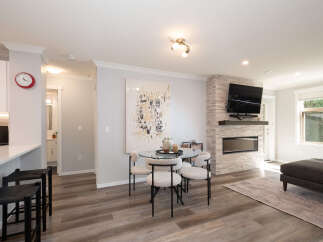 Experience modern living in this open-concept space featuring a sleek kitchen island with bar stools, a chic dining area with a glass table, and a cozy living room centered around a stone-clad fireplace with a mounted TV.