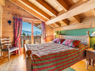Experience cozy mountain living in this charming chalet bedroom, featuring a comfortable bed adorned with festive linens and a stunning view of snow-capped peaks from the balcony.