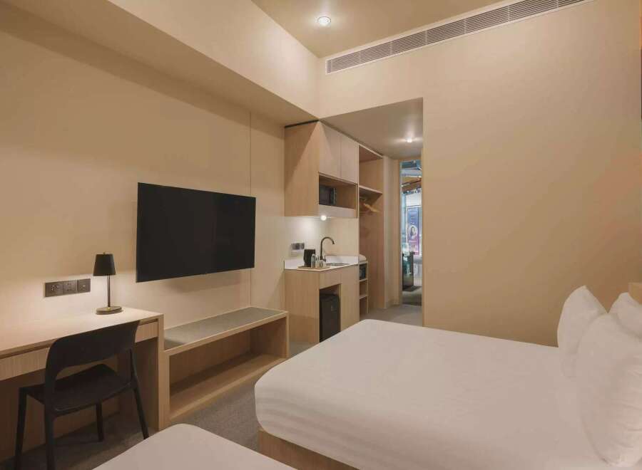 Experience modern comfort in this hotel room featuring two plush beds, a convenient kitchenette with a sink and mini-fridge, and a dedicated workspace with a desk and lamp.