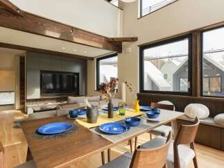 Experience modern living in this spacious dining and living area, featuring a large wooden dining table set for a meal and a comfortable sectional sofa facing a wall-mounted TV.