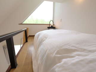 Experience cozy living in this charming loft bedroom, featuring a comfortable bed and a unique triangular window offering picturesque views of lush greenery.