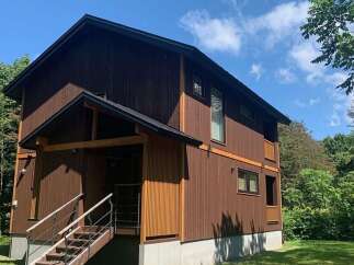 Discover this charming two-story wooden house nestled amidst lush greenery and a clear blue sky.