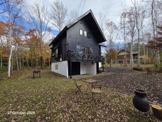 Experience the charm of autumn at this unique property, featuring a modern black house with a striking white foundation and a wooden deck.