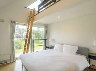 Experience modern living in this stylish bedroom featuring a comfortable bed, a loft accessible by a wooden ladder, and large windows offering scenic views.