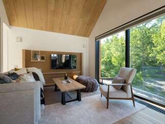 Experience modern living in this stunning living room, featuring a vaulted wood-paneled ceiling and a wall of windows offering breathtaking forest views.