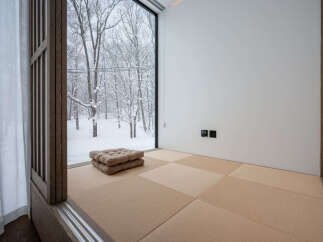 Experience serene winter living in this minimalist room featuring a large window with a picturesque snow-covered forest view.