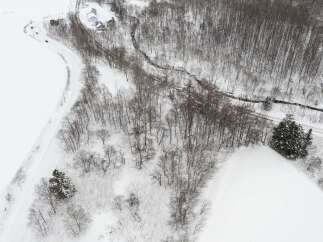 Discover serene winter living with this aerial view of a snow-covered landscape featuring a winding road and a charming cluster of houses nestled amongst bare trees.