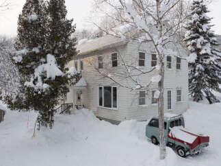Experience the charm of this winter wonderland property, featuring a classic white house nestled amidst snow-covered trees.