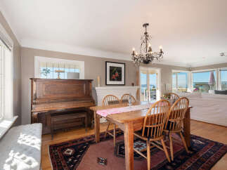 This spacious dining and living area boasts a beautiful antique piano, a charming wooden dining table with spindle-back chairs, and a cozy tufted bench by the window.