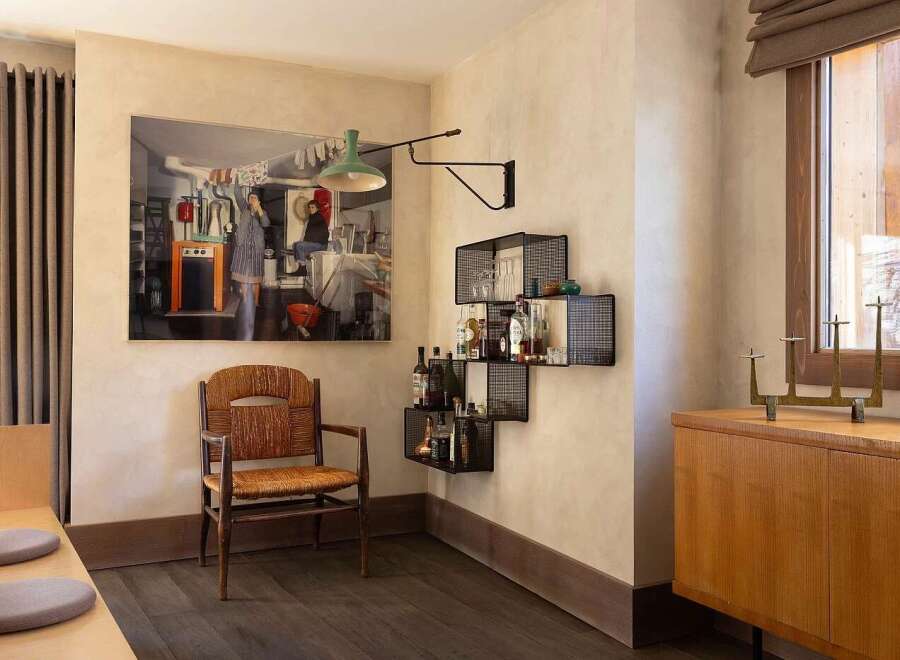 This inviting dining area features a large, captivating artwork depicting a domestic scene, complemented by a rustic wooden chair and a modern black metal bar shelf stocked with spirits and glassware.