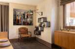 This inviting dining area features a large, captivating artwork depicting a domestic scene, complemented by a rustic wooden chair and a modern black metal bar shelf stocked with spirits and glassware.