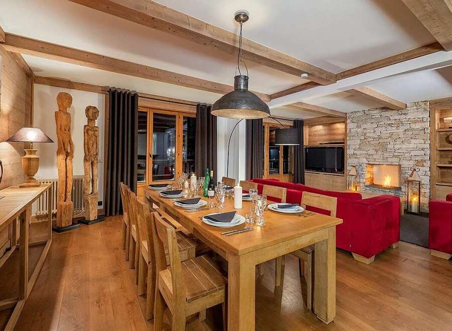 Experience luxurious alpine living in this stunning dining and living area, featuring a grand wooden dining table set for a feast and plush red seating arranged around a cozy stone fireplace.