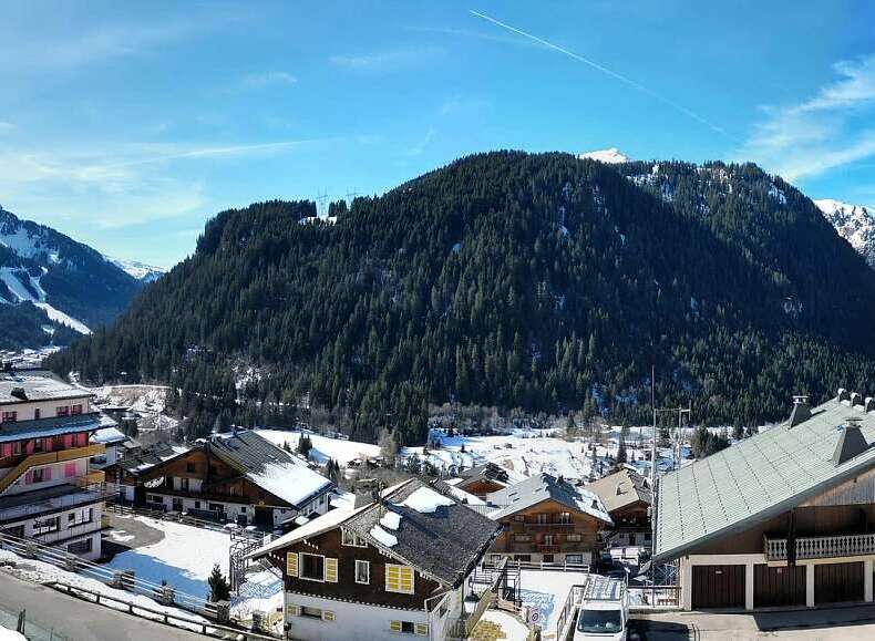 Experience the charm of a winter wonderland with this stunning panoramic view of a snow-covered village nestled at the foot of majestic, tree-lined mountains.