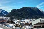 Experience the charm of a winter wonderland with this stunning panoramic view of a snow-covered village nestled at the foot of majestic, tree-lined mountains.
