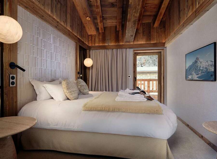 Experience luxury mountain living in this beautifully appointed bedroom, featuring a plush king-sized bed, warm wood accents, and a stunning view of snow-capped peaks through the balcony door.